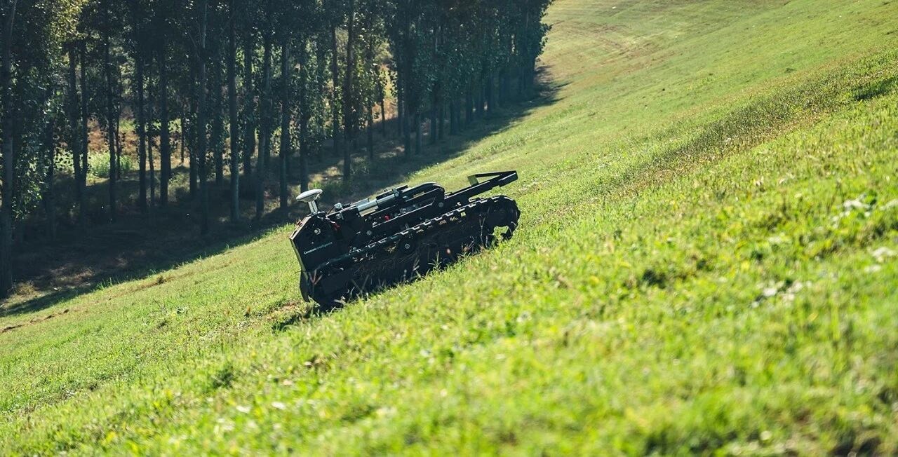 Tracked WANJI T1-900A autonomous flail mower working across a steep grassy embankment beside a tree line