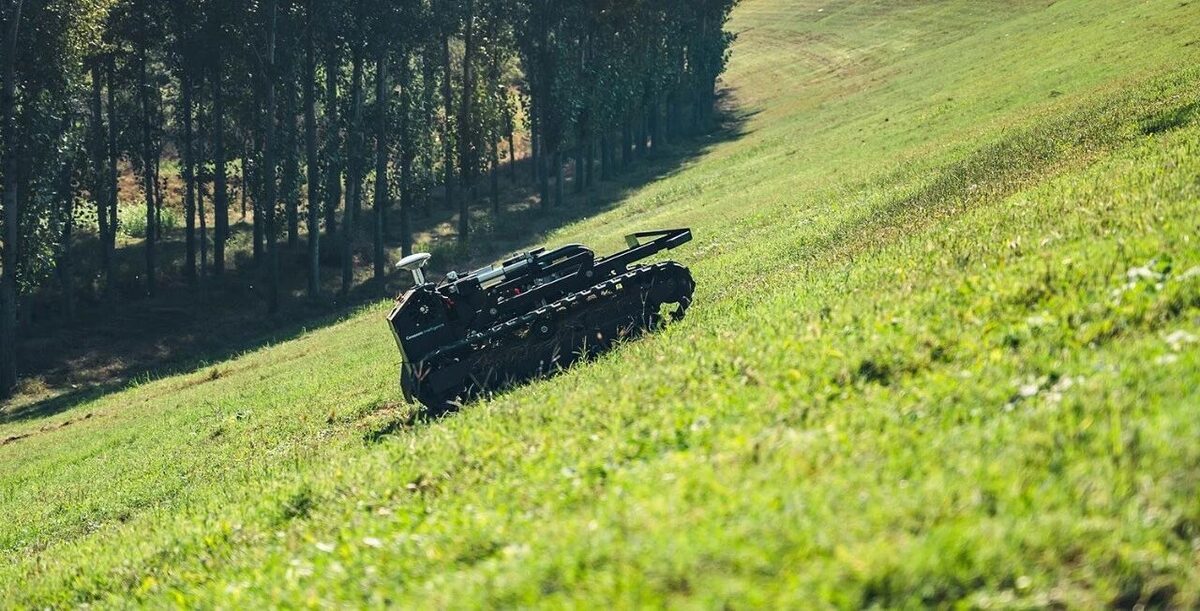 Tracked autonomous mower working across a steep green slope beside a tree line